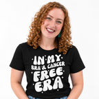 A smiling woman with curly red hair wearing a black graphic t-shirt that reads 'IN MY BRA' & CANCER FREE ERA' in bold white letters, set against a plain white background.