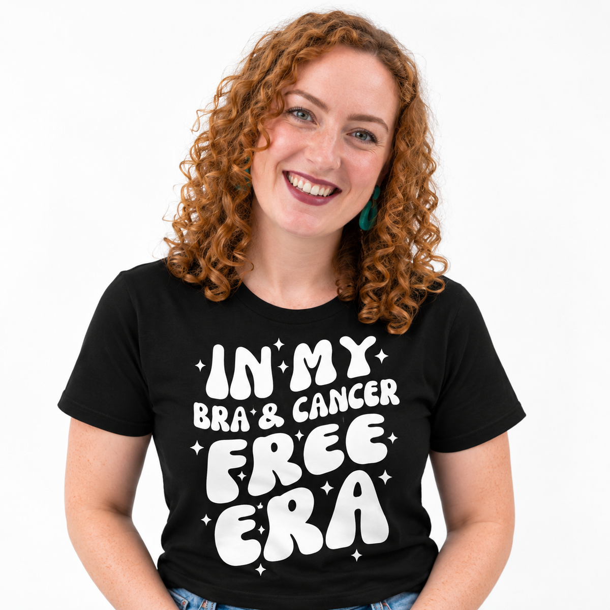 A smiling woman with curly red hair wearing a black graphic t-shirt that reads 'IN MY BRA' & CANCER FREE ERA' in bold white letters, set against a plain white background.