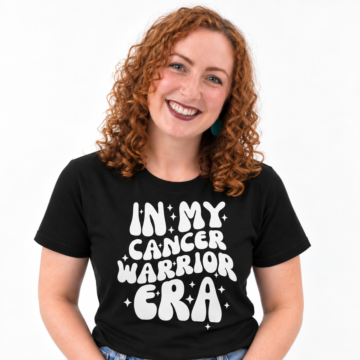 A smiling woman with curly red hair wearing a black short-sleeve T-shirt with the phrase 'IN MY CANCER WARRIOR ERA' printed in large white retro-style letters with small star accents, against a plain white background.