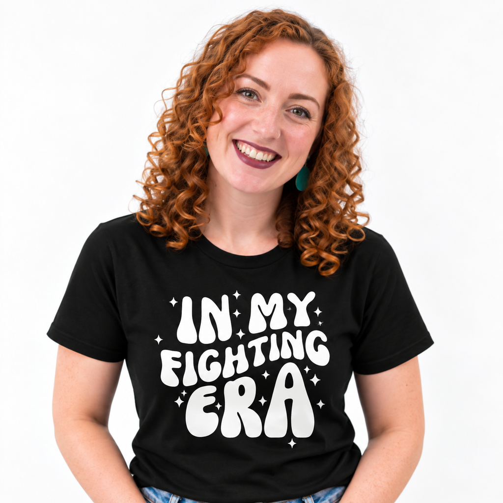 A smiling woman with curly red hair wearing a black graphic t-shirt that reads 'IN MY FIGHTING ERA' in white retro lettering with small white stars, set against a plain white background.