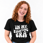 A smiling woman with curly red hair wearing a black graphic t-shirt that reads 'IN MY FIGHTING ERA' in white retro lettering with small white stars, set against a plain white background.