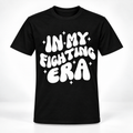 A black short-sleeve crewneck t-shirt with a bold white graphic text reading 'IN MY FIGHTING ERA' surrounded by small white star sparkles, displayed on a clean white background.