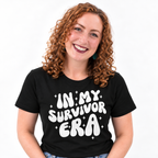 A smiling woman with curly red hair wearing a black t-shirt with the phrase 'IN MY SURVIVOR ERA' printed in a bold white retro font with small star accents, set against a plain white background.
