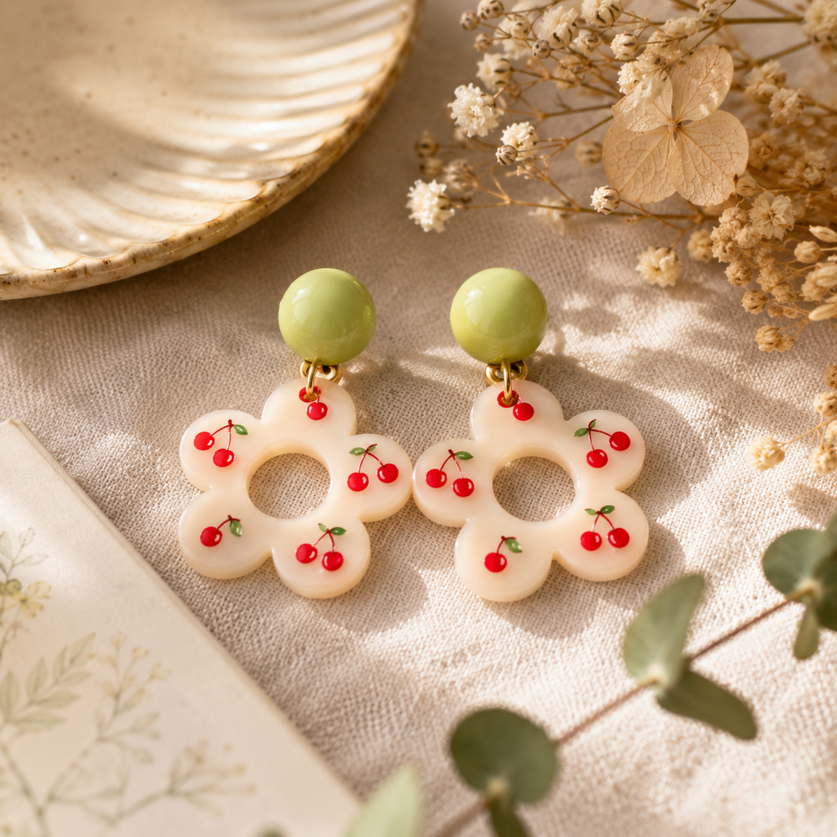 Cherry Flower Earrings with pink floral drops, red cherry details and glossy pale green round studs on woven table cloth surrounded by flowers and leaves