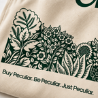 Close-up of a cream canvas tote bag featuring a dark green botanical floral pattern and the printed text 'Buy Peculiar. Be Peculiar. Just Peculiar.