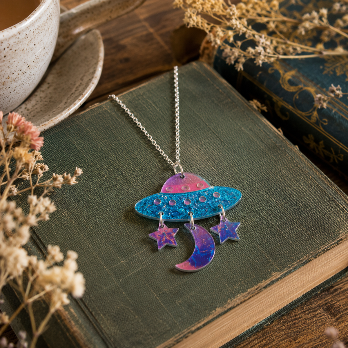Glitter UFO Necklace with blue spaceship pendant, pink dome and dangling purple crescent moon and star charms on a book