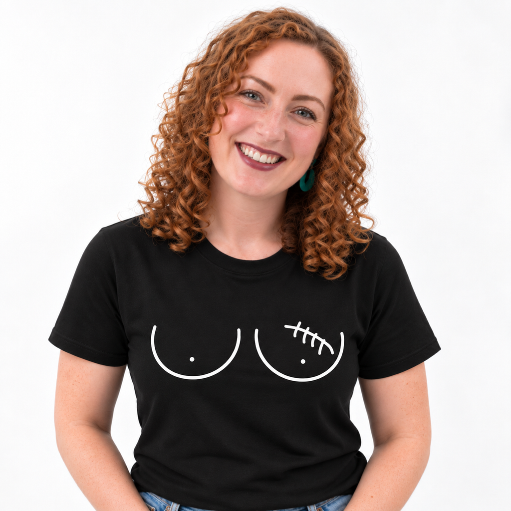 A smiling woman with curly red hair wearing a black short-sleeve t-shirt with a white graphic of two breasts, the left one with a lumpectomy scar, against a plain white background.