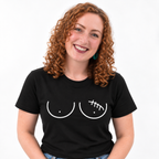 A smiling woman with curly red hair wearing a black short-sleeve t-shirt with a white graphic of two breasts, the left one with a lumpectomy scar, against a plain white background.