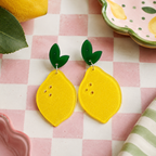 Lemon Dangle Earrings with bright yellow acrylic lemon drops and glossy green leaf-shaped studs on a pink and white checkered table