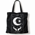 A black canvas tote bag with a white tarot card graphic of 'The Moon' featuring a crescent moon, stars, and hands, shown on a plain white background.