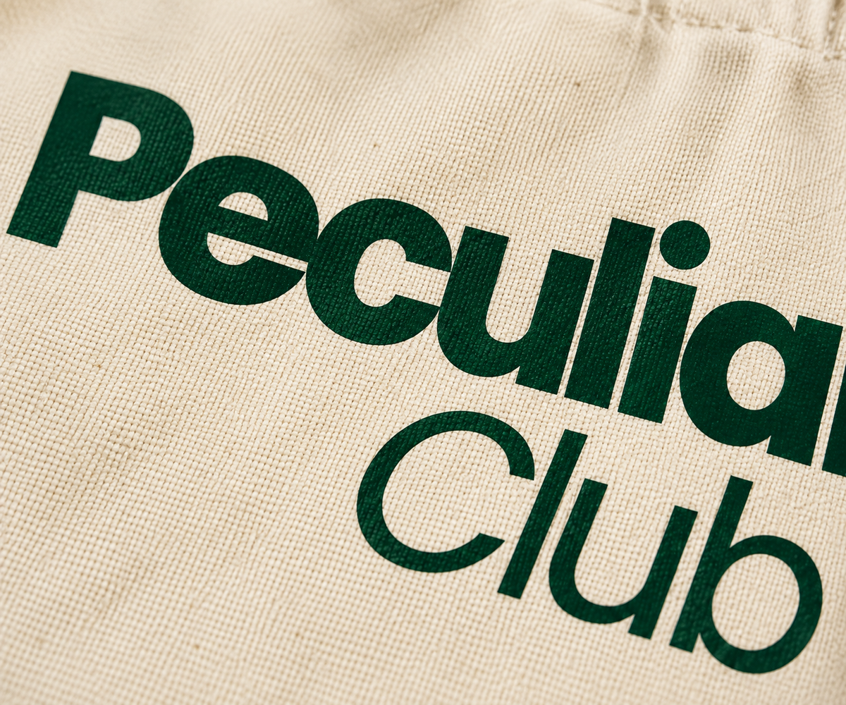 Close-up of a beige canvas tote bag printed with the words 'Peculiar Club' in bold dark green lettering.