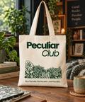 A cream canvas tote bag with green printed text reading 'Peculiar Club' and a botanical illustration, standing on a wooden table in a cozy, book-filled room.