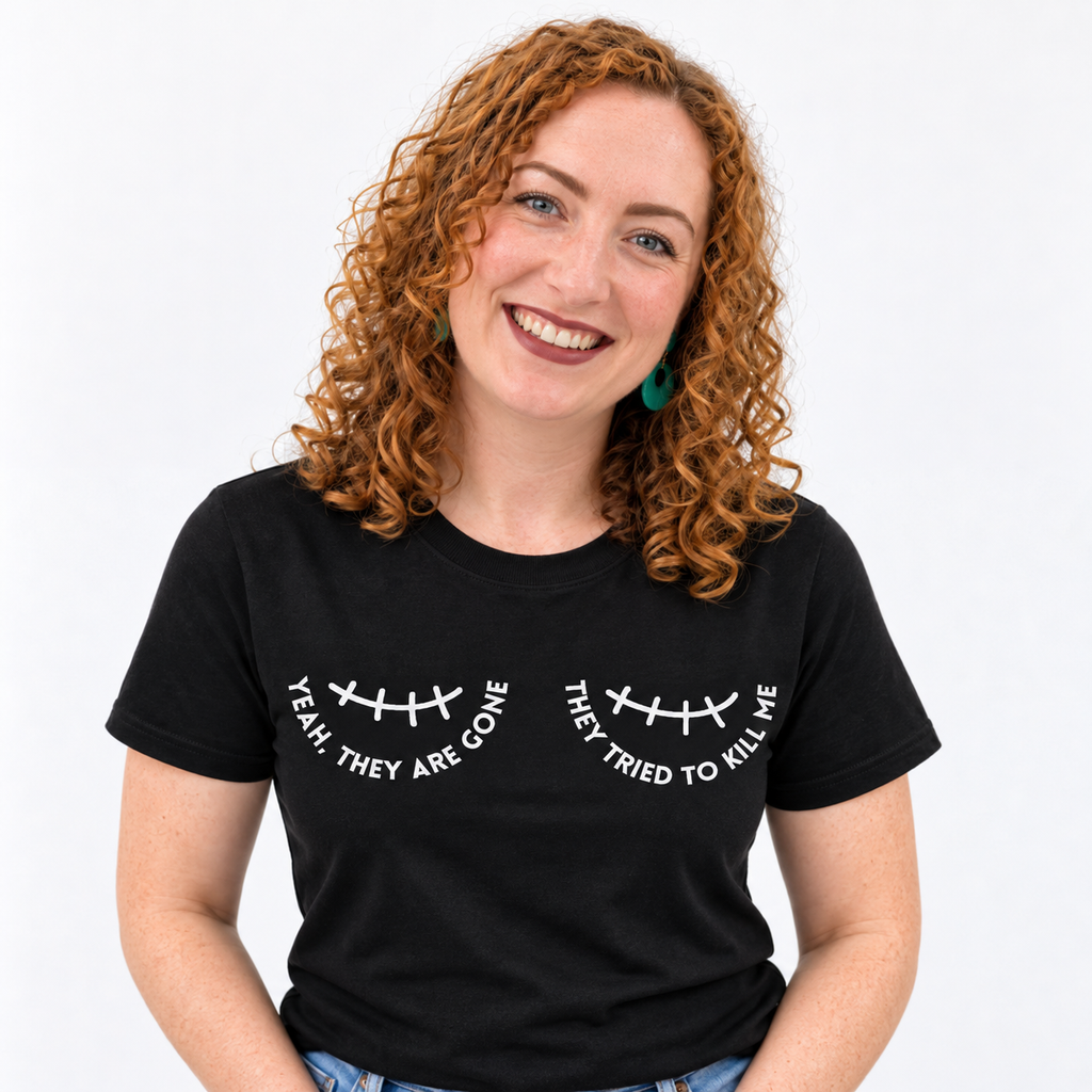 A smiling woman with curly red hair wearing a black graphic of a double mastectomy scar and the text 'YEAH THEY ARE GONE' and 'THEY TRIED TO KILL ME' printed on the chest.
