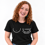 A smiling woman with curly red hair wearing a black short-sleeve t-shirt with a white graphic of a right breast and a left mastectomy scar and the text 'YEAH, THIS ONE'S GONE IT TRIED TO KILL ME' printed on the chest.