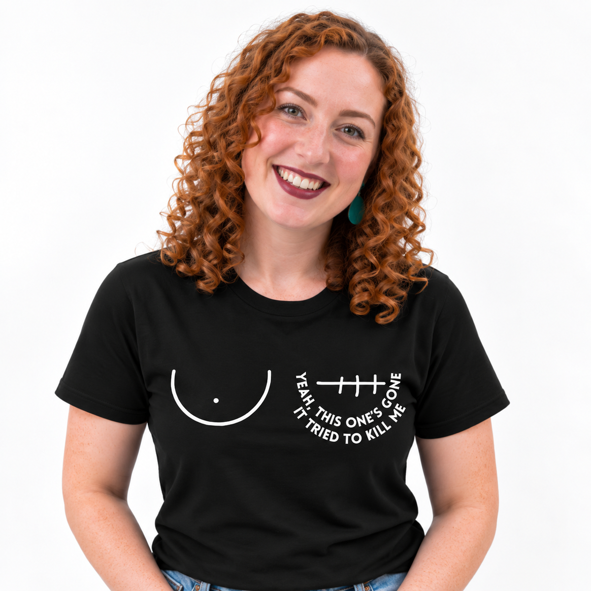 A smiling woman with curly red hair wearing a black short-sleeve t-shirt with a white graphic of a right breast and a left mastectomy scar and the text 'YEAH, THIS ONE'S GONE IT TRIED TO KILL ME' printed on the chest.