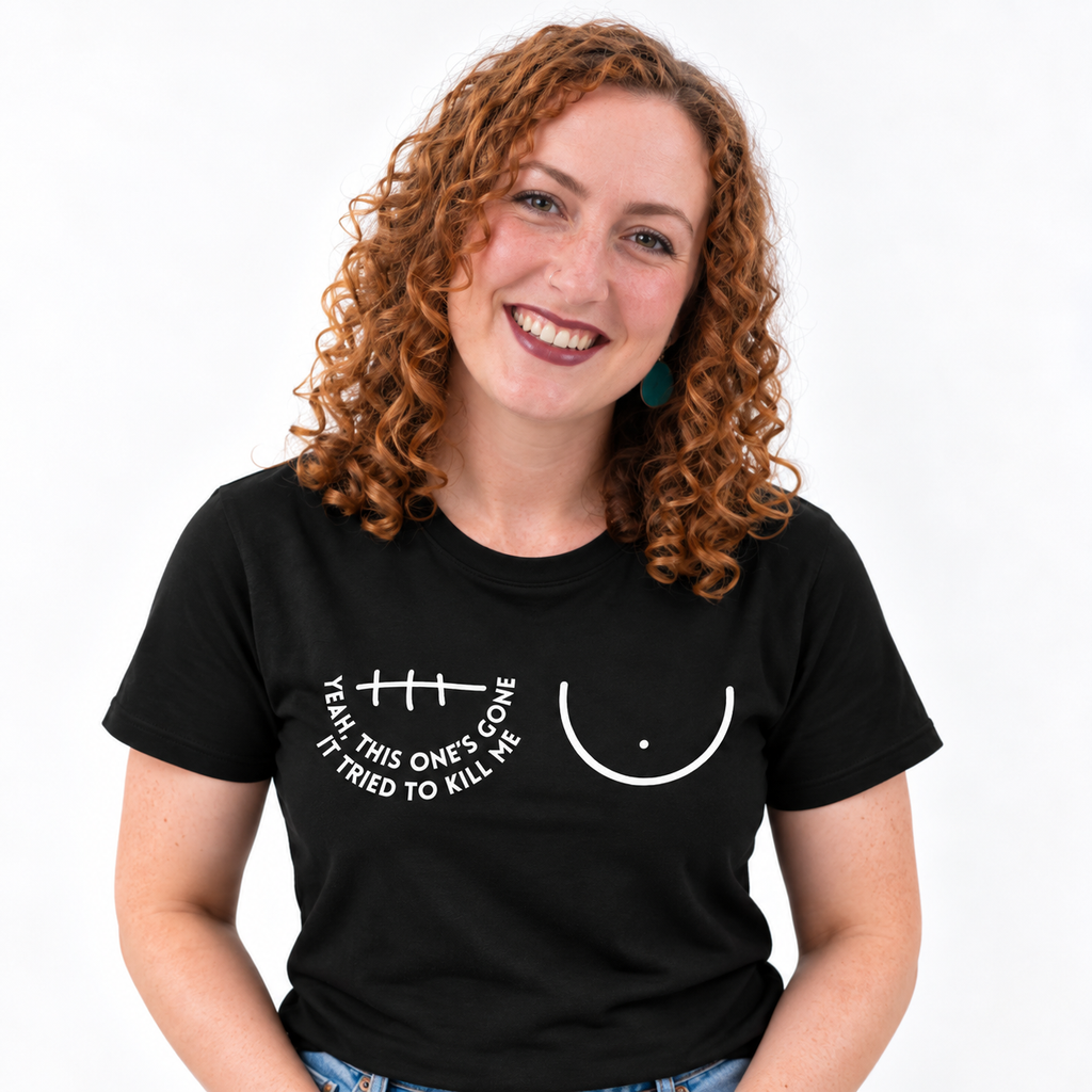 A smiling woman with curly red hair wearing a black short-sleeve t-shirt with a white graphic of a left breast and a right mastectomy scar and the text 'YEAH, THIS ONE'S GONE IT TRIED TO KILL ME' printed on the chest.