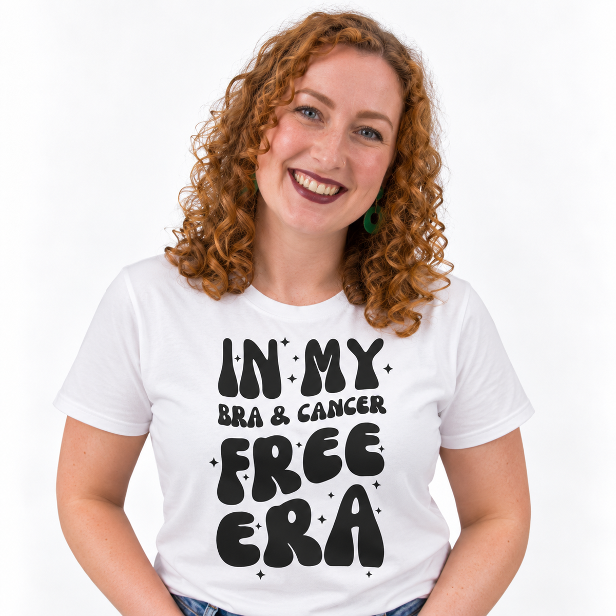 A smiling woman with curly red hair wearing a white graphic t-shirt that reads 'IN MY BRA & CANCER FREE ERA' in black retro lettering, set against a plain white background.