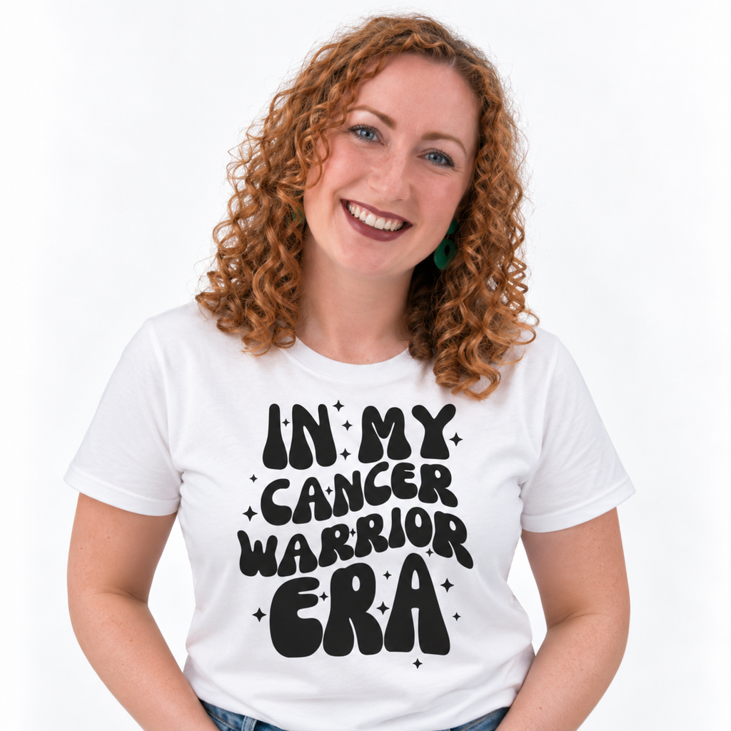 A smiling woman with curly red hair wearing a white graphic t-shirt with the text 'IN MY CANCER WARRIOR ERA' in black retro-style lettering, set against a plain white background.