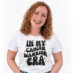 A smiling woman with curly red hair wearing a white graphic t-shirt with the text 'IN MY CANCER WARRIOR ERA' in black retro-style lettering, set against a plain white background.