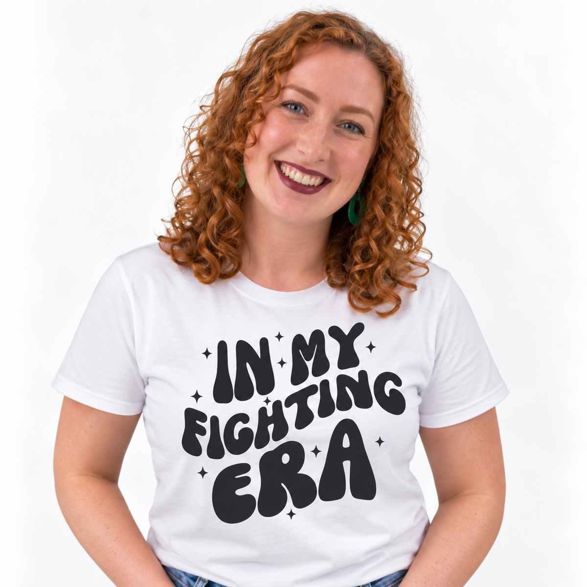 A smiling woman with curly red hair wearing a white graphic T-shirt with the text 'IN MY FIGHTING ERA' in bold black lettering, set against a plain white background.