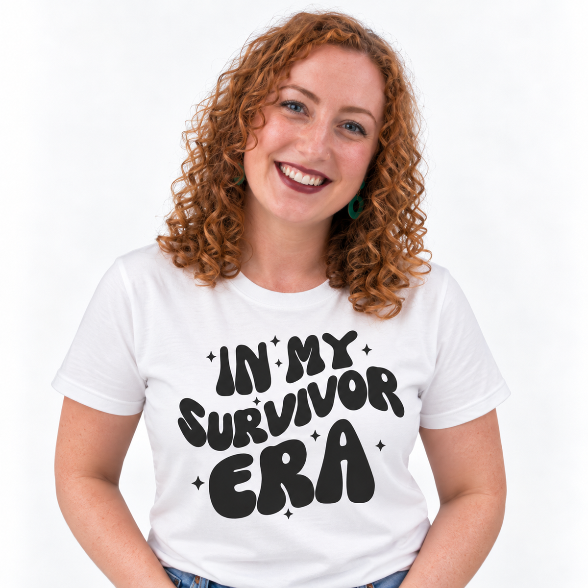 A smiling woman with curly red hair wearing a white graphic T-shirt with the text 'IN MY SURVIVOR ERA' in black retro lettering, set against a plain white background.